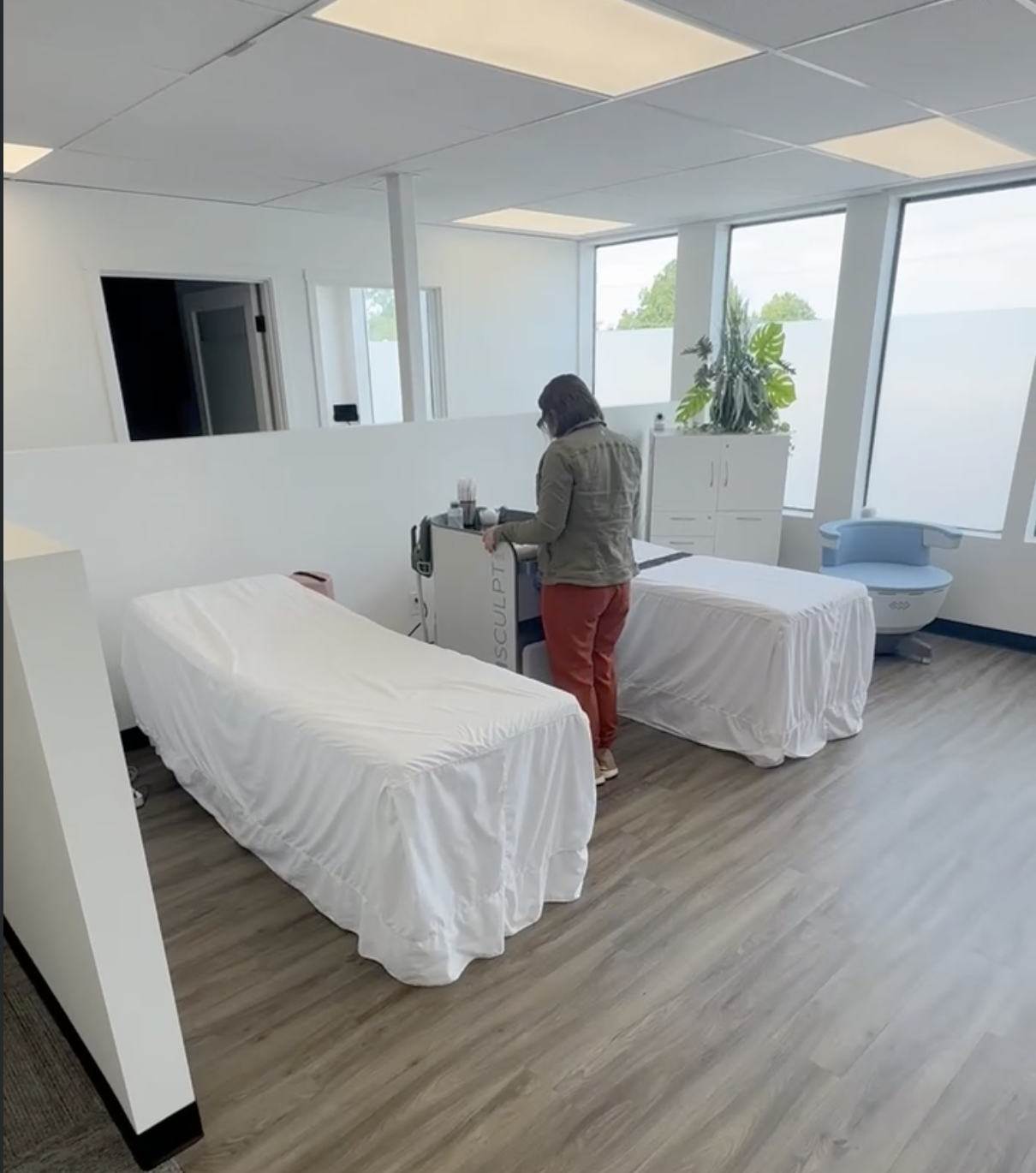 Renee Storm setting up an EMSCULPT device between two treatment tables in a bright, window-lined treatment room